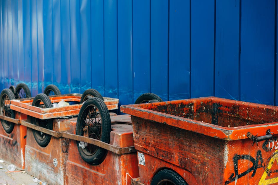 A row of large orange skips with black rubber wheels, positioned against a tall blue metal fence, used for disposal of bulky waste during house removals. The skips are made of reinforced plastic and metal, with some showing signs of dirt and heavy use. They are placed on a paved outdoor surface, with one skip partially filled with debris and covered in dirt. The other skip is empty, ready for collection. The background features vertical panels of the blue fence, indicating an outdoor area likely associated with a moving or disposal service. The scene illustrates the process of waste management in connection with home relocation, facilitated by Removal Companies Merton, for efficient packing and loading during removals.