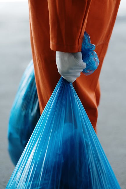 A row of large orange skips with black rubber wheels, positioned against a tall blue metal fence, used for disposal of bulky waste during house removals. The skips are made of reinforced plastic and metal, with some showing signs of dirt and heavy use. They are placed on a paved outdoor surface, with one skip partially filled with debris and covered in dirt. The other skip is empty, ready for collection. The background features vertical panels of the blue fence, indicating an outdoor area likely associated with a moving or disposal service. The scene illustrates the process of waste management in connection with home relocation, facilitated by Removal Companies Merton, for efficient packing and loading during removals.