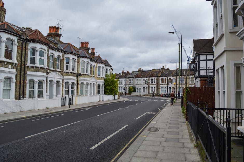 A quiet residential street in Colliers Wood with traditional Victorian-style terraced houses featuring white-painted facades, bay windows, and red brick chimneys. The pavement runs alongside the row of houses, separated by black metal railings, and is lined with modern street lamps and a few small trees. The street is empty except for a single person walking away in the distance, and several parked cars are visible along the curb. Overhead, a cloudy sky casts a diffuse light over the scene. This setting illustrates typical environments where house removals and furniture transport services by Removal Companies Merton may take place during local property moves, with visible elements such as the street layout, residential facades, and parking arrangements supporting an understanding of moving logistics in this neighbourhood.