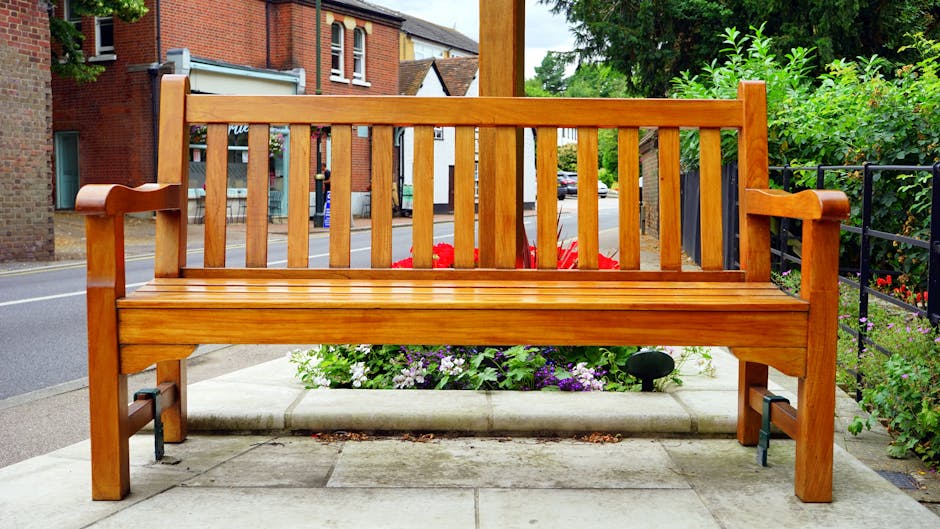 A polished wooden park bench with armrests and slatted backrest, positioned on a concrete sidewalk in a residential area, with a flower bed containing purple and red flowers directly behind it. The background shows brick houses with large windows, a street with parked cars, and green trees and shrubs providing greenery. To the right, a black metal railing borders the sidewalk, and part of a black truck or van is visible in the distance. The scene is well-lit, suggesting daytime, and the setting appears peaceful, suitable for a home relocation or moving context as part of a quiet suburban street, with the bench potentially used as a resting spot during furniture transport or packing activities, as exemplified by removals services like Removal Companies Merton.