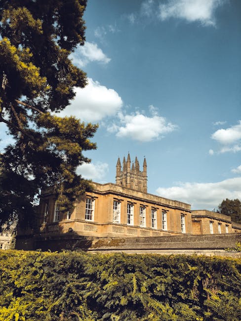 A historic stone building with multiple large, rectangular windows and a prominent central tower featuring pointed pinnacles, situated on a landscaped area with low greenery and trees. The building is illuminated by natural daylight, with a partly cloudy sky overhead. In the foreground, a large evergreen tree partially obscures the view of the structure. The scene depicts a calm, outdoor setting, possibly part of a countryside estate or university campus area, which Removal Companies Merton may service for house relocations involving historical properties or estate moves, showcasing the importance of careful furniture transport and packing during a home relocation process.