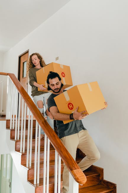 Two movers from Removal Companies Merton are carrying large cardboard boxes with red 'H' symbols down a wooden staircase inside a home, with a woman guiding them from the upper level. The boxes are secured with packing tape and are being carefully handled during the home relocation process. The staircase features wooden steps and a wooden handrail, with a plain white wall in the background. The natural lighting illuminates the indoor environment, emphasizing the careful loading process that is part of packing and moving services. The scene reflects the detailed logistics involved in furniture transport and home packing for a local move in Merton, with movers carefully managing their load through internal stairs as part of the overall removal service.