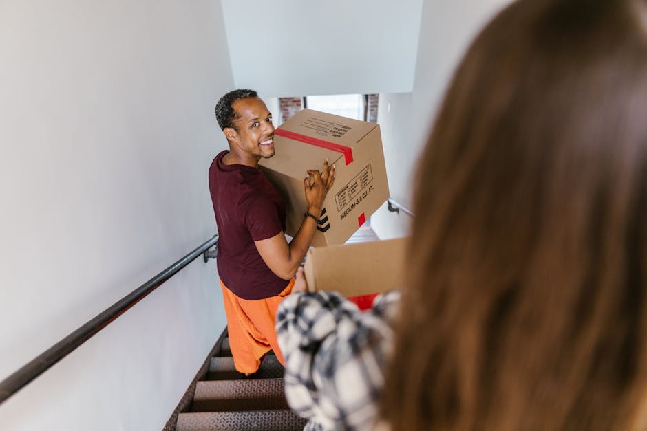 A man is seen on a staircase inside a home, smiling while holding a medium-sized cardboard box sealed with red tape and labeled with handling instructions. He is wearing a maroon t-shirt and orange pants, indicating he is engaged in home relocation or furniture transport as part of a removals process. In the foreground, a second person with long brown hair, wearing a checkered shirt, is assisting by holding another box, suggesting teamwork during packing or loading. The staircase has a black handrail attached to the white wall, and natural light filters in from a window above, illuminating the scene. This setting reflects the logistics of packing and moving household items, a typical part of professional removals by companies like Removal Companies Merton, especially related to budgeting for additional costs such as stairs or packing materials amid house removals in Merton.
