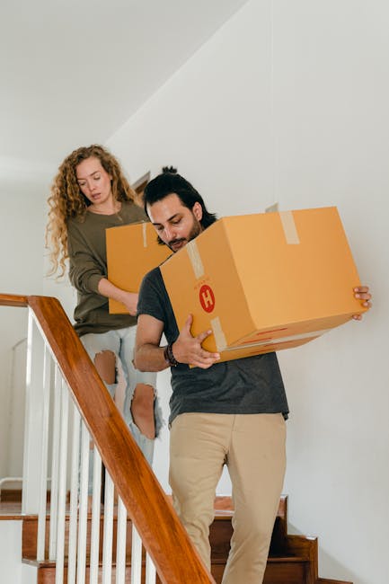Two movers from Removal Companies Merton are carrying large cardboard boxes with red 'H' symbols down a wooden staircase inside a home, with a woman guiding them from the upper level. The boxes are secured with packing tape and are being carefully handled during the home relocation process. The staircase features wooden steps and a wooden handrail, with a plain white wall in the background. The natural lighting illuminates the indoor environment, emphasizing the careful loading process that is part of packing and moving services. The scene reflects the detailed logistics involved in furniture transport and home packing for a local move in Merton, with movers carefully managing their load through internal stairs as part of the overall removal service.