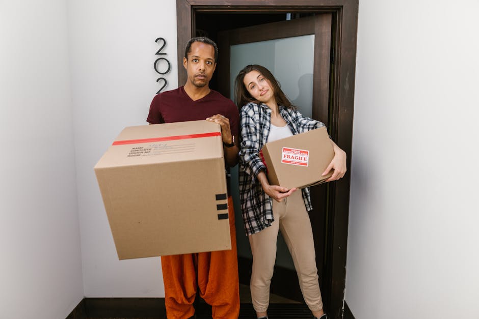 A man and a woman stand inside a residential hallway, near a doorway, holding cardboard boxes filled with belongings, as part of a home relocation process. The man, dressed in a maroon t-shirt and orange pants, is holding a large box with black tape, while the woman, wearing a plaid shirt and beige trousers, carries a smaller box labeled 'FRAGILE' in red. Behind them, the doorway leads to another room, with a dark-framed glass door visible. The hallway has plain white walls, with the number '200' displayed vertically beside the door frame, and is lit with natural or neutral indoor lighting. The scene depicts a loading or packing step during a house move, with visible packaging materials such as cardboard boxes, and suggests a professional removal or moving service like Removal Companies Merton assisting with a same-day or late notice move, emphasizing furniture transport and packing and moving logistics.