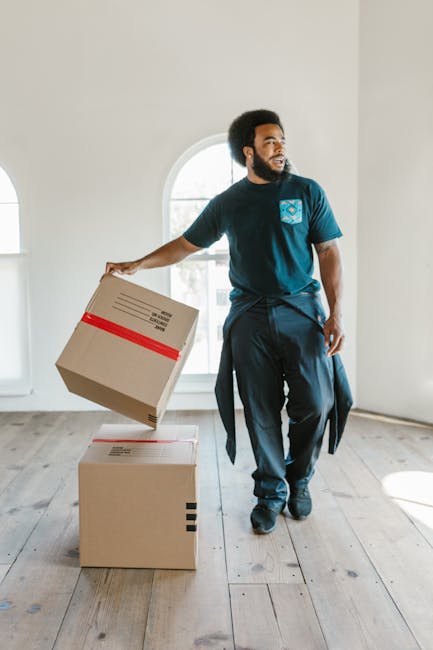 A man with dark curly hair and a beard, dressed in a navy T-shirt with a logo, dark pants, and black shoes, stands inside a spacious room with white walls and large arched windows that allow natural light to fill the space. He is holding a cardboard moving box with red packing tape in his right hand, which he appears to be preparing to carry or move, and there are two additional cardboard boxes stacked on the wooden floor next to him. The room is empty of furniture, and the flooring is light-colored wood. This scene depicts the initial stages of a home relocation or packing process, which might be part of a same-day or late notice move handled by a professional moving service such as Removal Companies Merton, with the focus on packing, handling, or organizing household items for transport.