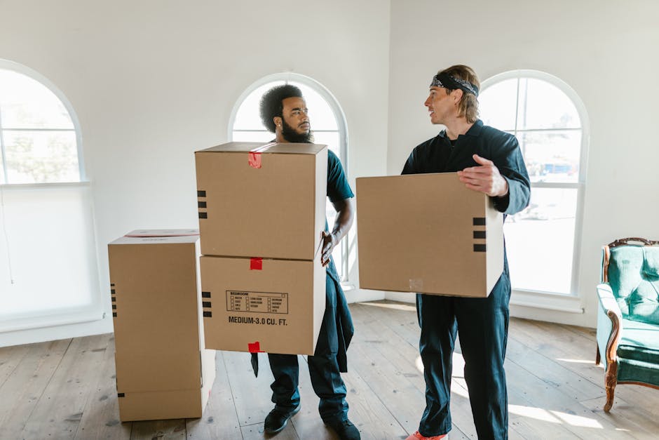 A man and a woman stand inside a residential hallway, near a doorway, holding cardboard boxes filled with belongings, as part of a home relocation process. The man, dressed in a maroon t-shirt and orange pants, is holding a large box with black tape, while the woman, wearing a plaid shirt and beige trousers, carries a smaller box labeled 'FRAGILE' in red. Behind them, the doorway leads to another room, with a dark-framed glass door visible. The hallway has plain white walls, with the number '200' displayed vertically beside the door frame, and is lit with natural or neutral indoor lighting. The scene depicts a loading or packing step during a house move, with visible packaging materials such as cardboard boxes, and suggests a professional removal or moving service like Removal Companies Merton assisting with a same-day or late notice move, emphasizing furniture transport and packing and moving logistics.
