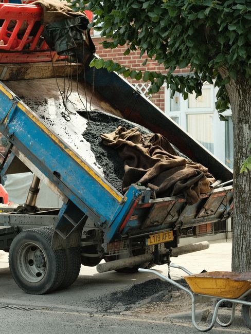 A small blue and yellow dump truck with an open bed is parked on a residential street near a brick house and a tree. The truck's bed contains dark asphalt, a brown cloth, and some packaging materials, indicating ongoing road or driveway repairs related to house removals or moving logistics. A wheelbarrow with a yellow basin is positioned on the pavement next to the truck, and a tree with green leaves partially obscures the scene. The truck's rear license plate reads L119 AFE. The setting suggests a street-loading activity or preparation work for home relocation, with attention to safety and proper equipment usage, as seen in the presence of the truck and wheelbarrow, supporting professional removals or packing and moving services like those offered by Removal Companies Merton.