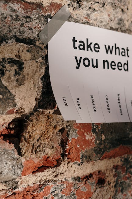 A close-up image of a white paper with the heading 'take what you need' taped to a rustic brick wall. The paper features several words listed vertically including 'love', 'hope', 'passion', 'courage', and 'happiness', suggesting a motivational message. The background consists of exposed, weathered bricks with varying shades of red, brown, and gray, and the paper is affixed with a piece of tape at the top. This visual is relevant to the context of house removals and moving services, reflecting themes of support and positivity during home relocation, as provided by Removal Companies Merton.