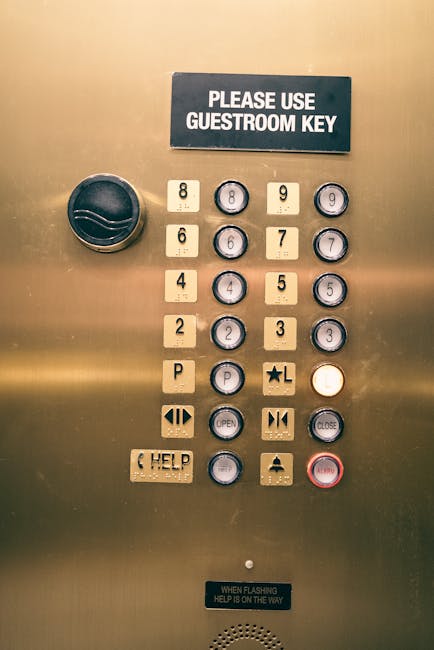 A close-up view of an elevator control panel with a metallic gold surface, featuring a rectangular black sign at the top that reads 'PLEASE USE GUESTROOM KEY' in white text. The panel includes multiple round and rectangular buttons, with the floor buttons numbered 2 through 9 and 4, 6, and 8 displayed, alongside a 'P' for parking. There are additional function buttons such as 'OPEN' and 'CLOSE', with some buttons marked with symbols like arrows and a star. A circular black knob is located on the left side of the panel, and a small rectangular button with a help icon and text 'HELP' is near the bottom. The panel also has a small white sign at the bottom stating, 'WHEN FLASHING HELP IS ON THE WAY'. This control panel is typically found in residential buildings or hotels, used for stair access, luggage transport, and elevator operation, and forms part of the logistics involved in house removals. The context suggests its role in facilitating apartment or flat relocations, supported by professional removal services such as those provided by Removal Companies Merton.
