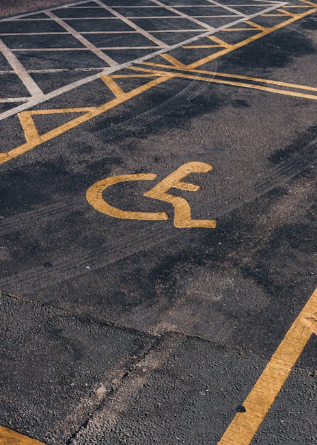 A close-up view of an asphalt parking area featuring a yellow wheelchair accessibility symbol painted on the ground, alongside yellow striped lines designating parking spaces and access areas. The surface shows signs of wear and slight cracking. The image emphasizes the outdoor environment, likely near a residential property or street where house removals or moving services might take place. Such parking markings are relevant for planning vehicle access during home relocation, as demonstrated by Removal Companies Merton on their website, which offers professional removals and packing services near Wimbledon Common, ensuring smooth furniture transport and efficient loading processes.
