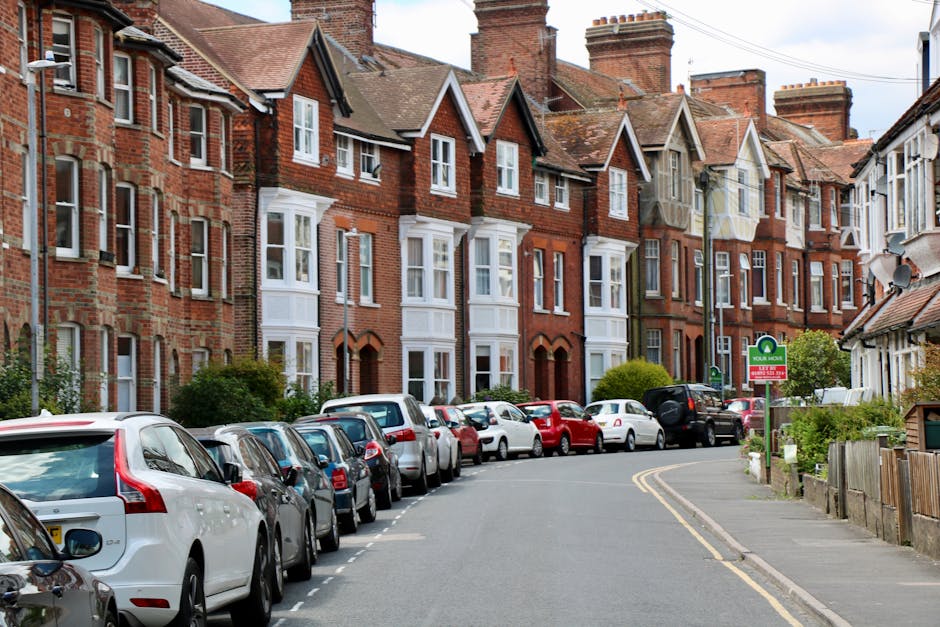 A residential street in Merton featuring a row of red brick Victorian-style terraced houses with bay windows and tiled roofs, situated behind a line of parked cars including hatchbacks, sedans, and SUVs along the curb. The vehicles cover both sides of the narrow, curved asphalt road, which includes yellow lines indicating parking restrictions in certain areas. sidewalk pavement runs alongside the parked vehicles, with a small green street sign visible on a pole indicating designated parking zones. A few bushes and small trees are planted in front of some houses, and a satellite dish is mounted on one roof. The scene is captured during daylight with a partly cloudy sky overhead, illustrating the typical urban environment where home relocations and furniture transport may involve parking considerations, as highlighted during discussions of parking suspension fines for moving activities, supported by Removal Companies Merton.