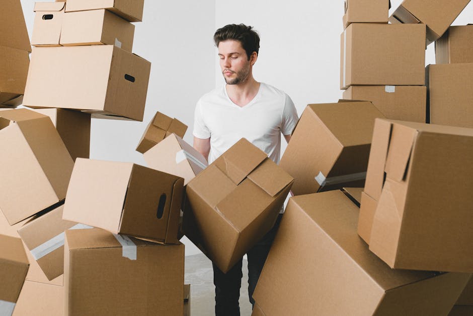 A man with dark hair and a short beard, dressed in a white t-shirt, standing indoors surrounded by numerous stacked cardboard boxes of various sizes, some sealed with white packing tape. The boxes are placed on the floor and appear to be part of a home relocation or packing process, with some boxes in mid-air, indicating they are being lifted or moved. The environment is well-lit with neutral-toned walls, suggesting a clean, prepared space for a moving or storage activity. This scene reflects the packing and moving stage often handled by professional removals services like Removal Companies Merton, involving careful handling of household items during a house move or storage solution.