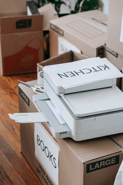 A cardboard moving box labeled 'BOOKS' positioned on a wooden floor, with a white multifunction printer placed on top of another box. The scene shows various packed boxes around, including a large box labeled 'KITCHEN' and others in the background, some open and some sealed with packing tape. The setting appears to be inside a property, near an open doorway or hallway, during a home relocation process organized by Removal Companies Merton, with typical packing materials such as cardboard, plastic wrap, and paper visible, indicating preparation for furniture transport or storage after a Merton move.