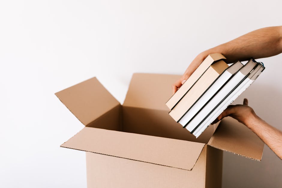 A man with dark hair and a short beard, dressed in a white t-shirt, standing indoors surrounded by numerous stacked cardboard boxes of various sizes, some sealed with white packing tape. The boxes are placed on the floor and appear to be part of a home relocation or packing process, with some boxes in mid-air, indicating they are being lifted or moved. The environment is well-lit with neutral-toned walls, suggesting a clean, prepared space for a moving or storage activity. This scene reflects the packing and moving stage often handled by professional removals services like Removal Companies Merton, involving careful handling of household items during a house move or storage solution.