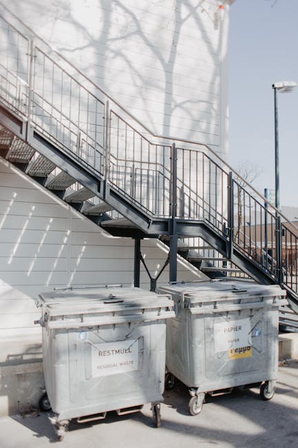 An outdoor scene showing two large grey wheeled waste disposal bins positioned on a paved area beneath a metal staircase with orange handrails, attached to the side of a building with white exterior walls. The bins are labeled for recycling waste and paper, respectively, with the paper bin marked 'RECYCLABLE PAPER' and 'PAPIER' in black text, and both have lids, handles, and are made of durable plastic. The staircase leads up to an upper level, casting shadows on the wall behind, and the area is well-lit by natural daylight. The scene relates to a home relocation or removal process, where waste collection and legal disposal notes might be involved, as seen on the bins, with [COMPANY_NAME] providing removal and moving services in Merton. The overall environment emphasizes organized waste management during packing or moving activities within a residential or commercial property setting.