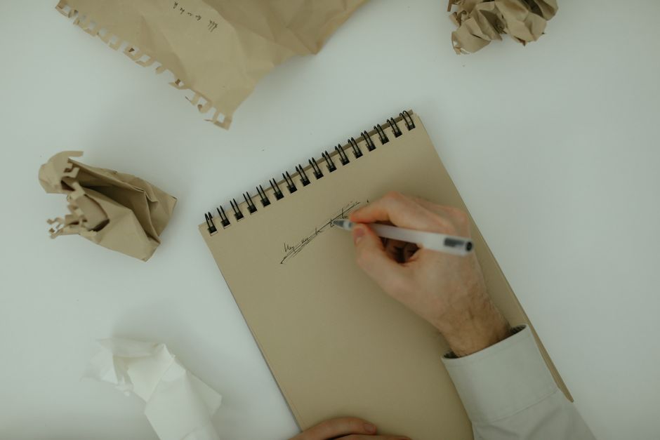 A person's hand, wearing a white shirt, is seen holding a black pen and signing a document on a small spiral-bound notepad placed on a white table. On the table, there are several crumpled pieces of brown and white paper, and a few sheets of beige packing paper with a perforated edge. In the background, a cardboard box with packing paper inside is partially visible, suggesting a packing or moving context. The scene appears to be indoors, with focus on the signing process related to home relocation, possibly for legal or disposal documentation as part of a house removals or moving service provided by Removal Companies Merton.