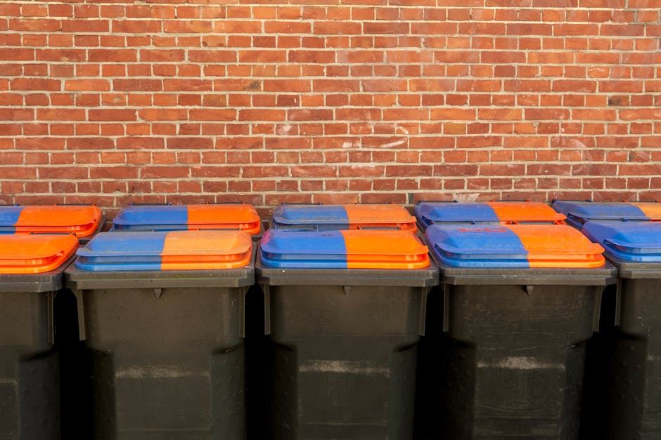An outdoor scene showing two large grey wheeled waste disposal bins positioned on a paved area beneath a metal staircase with orange handrails, attached to the side of a building with white exterior walls. The bins are labeled for recycling waste and paper, respectively, with the paper bin marked 'RECYCLABLE PAPER' and 'PAPIER' in black text, and both have lids, handles, and are made of durable plastic. The staircase leads up to an upper level, casting shadows on the wall behind, and the area is well-lit by natural daylight. The scene relates to a home relocation or removal process, where waste collection and legal disposal notes might be involved, as seen on the bins, with [COMPANY_NAME] providing removal and moving services in Merton. The overall environment emphasizes organized waste management during packing or moving activities within a residential or commercial property setting.